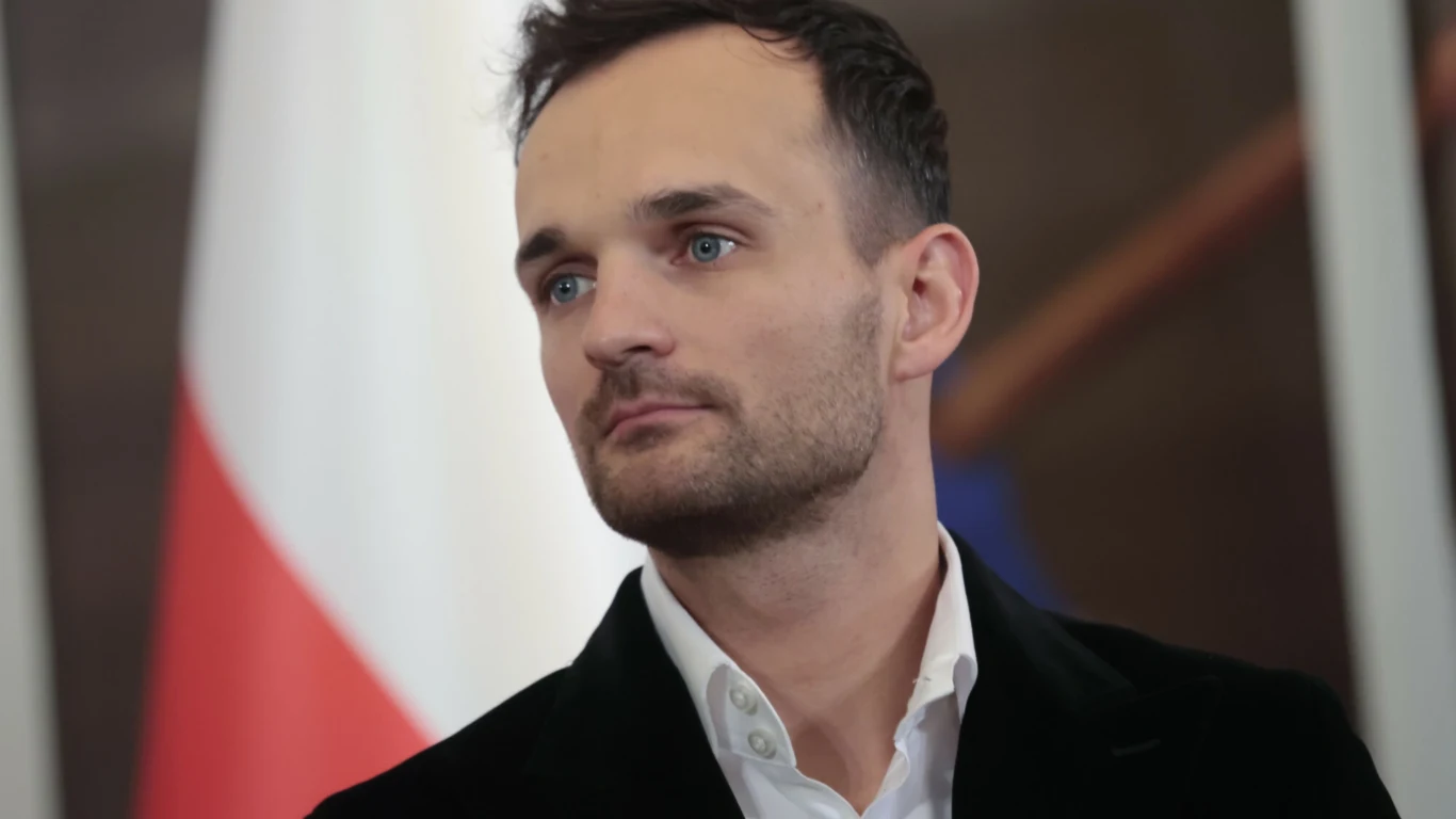 Mężczyzna o jasnych oczach i krótkich włosach w eleganckiej marynarce stoi na tle flagi Polski i nieostrego fragmentu niebieskiego elementu.