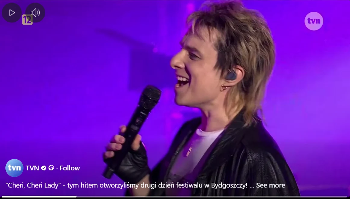 Wokalista z mikrofonem śpiewa na scenie, w tle intensywne fioletowe światło i lekko rozmyte tło, artysta ubrany w czarną kurtkę i rękawiczki.