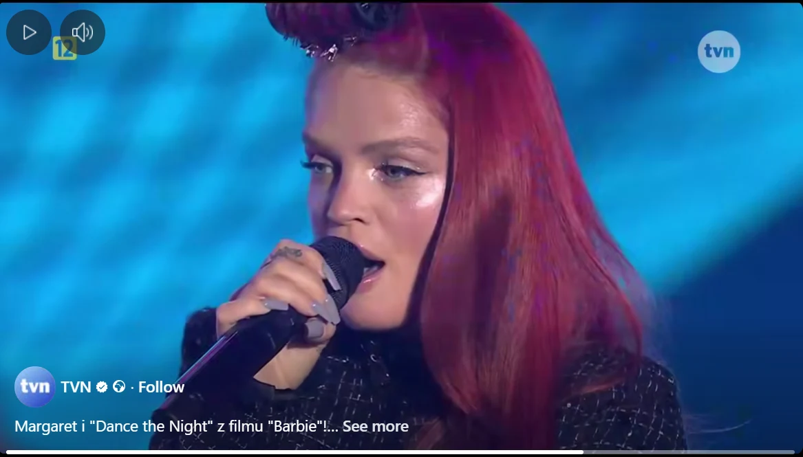 Kobieta z czerwonymi włosami śpiewa do mikrofonu na scenie, w tle niebieskie oświetlenie, na jej twarzy widoczny błyszczący makijaż.