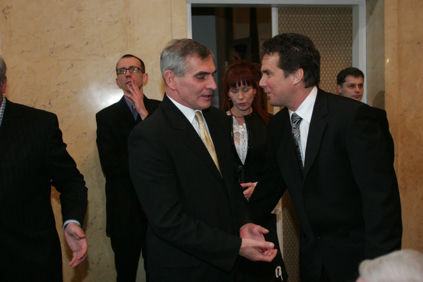 Kilku mężczyzn w garniturach rozmawia ze sobą w eleganckim, jasnym wnętrzu, w tle stoją inni uczestnicy spotkania, wszyscy zachowują poważny wyraz twarzy.