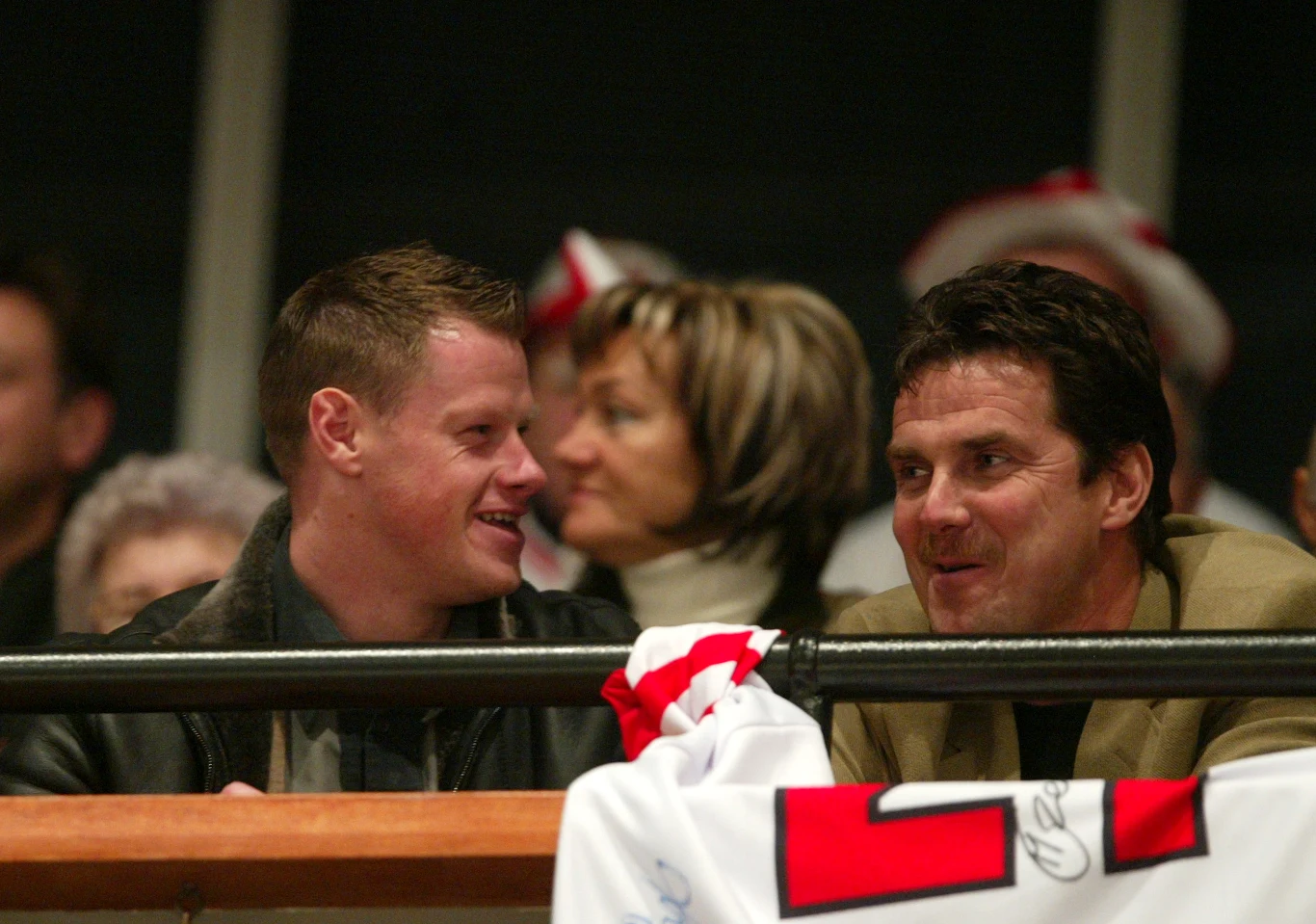 Dwóch mężczyzn siedzi obok siebie, rozmawiając i uśmiechając się. W tle widoczne są osoby kibicujące oraz fragment biało-czerwonej koszulki z numerem i podpisem na barierce.