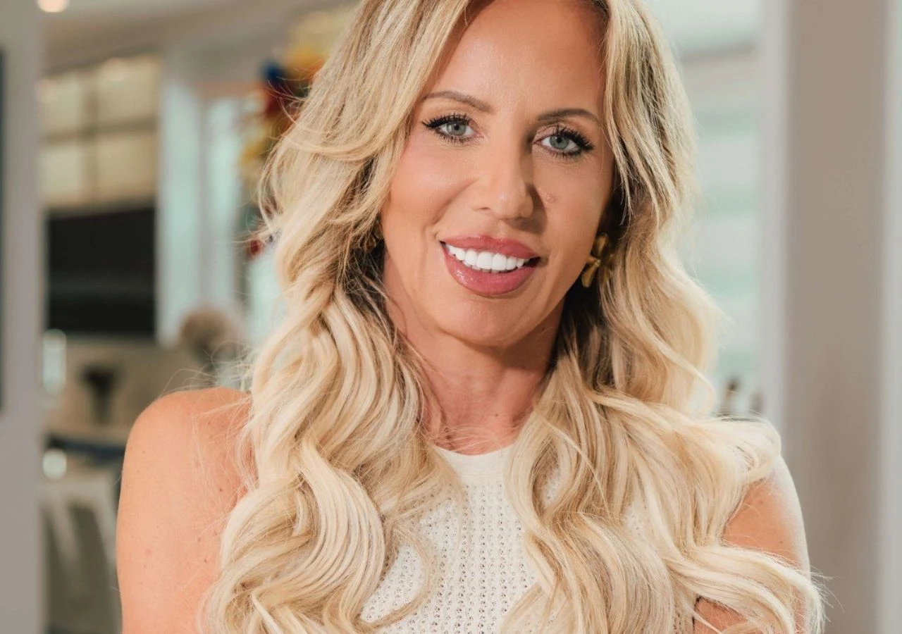 Dorota z "Żony dla Polaka" Kobieta o długich, blond włosach uśmiecha się, ubrana w jasny top. W tle rozmyte, nowoczesne wnętrze.