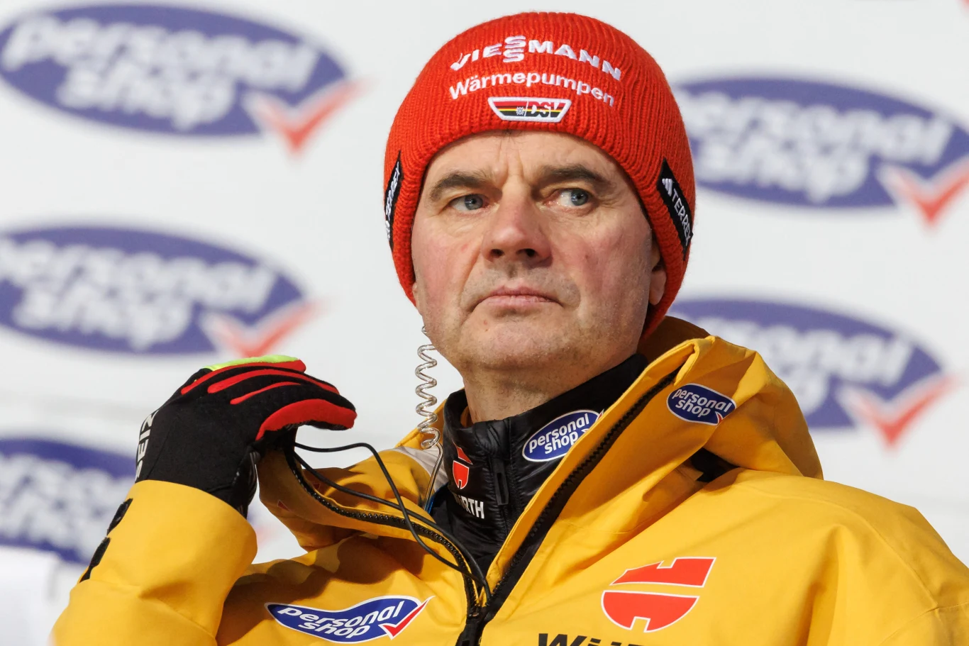 Stefan Horngacher jako szkoleniowiec reprezentacji Niemiec Mężczyzna w czerwonej czapce z logo i żółtej kurtce sportowej, z radiokomunikatorem w uchu, stoi przed tablicą z rozmazanym tłem zawierającym logotypy. Wygląda poważnie, patrzy lekko w bok.