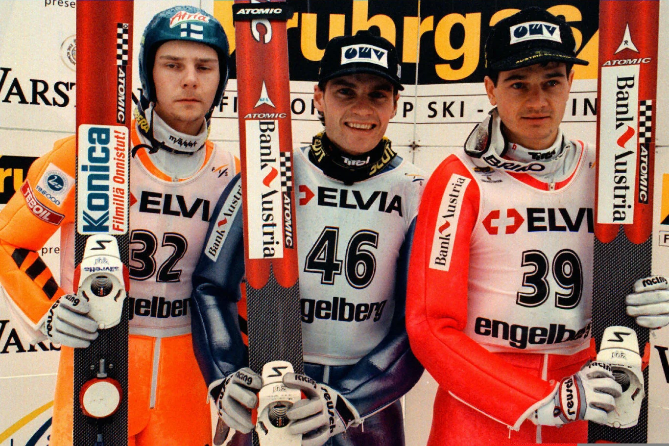 Trzech skoczków narciarskich w strojach sportowych trzyma narty, stojąc na podium z widocznymi numerami startowymi oraz logotypami sponsorów. Za nimi widoczne są reklamy oraz tło charakterystyczne dla zawodów sportowych.