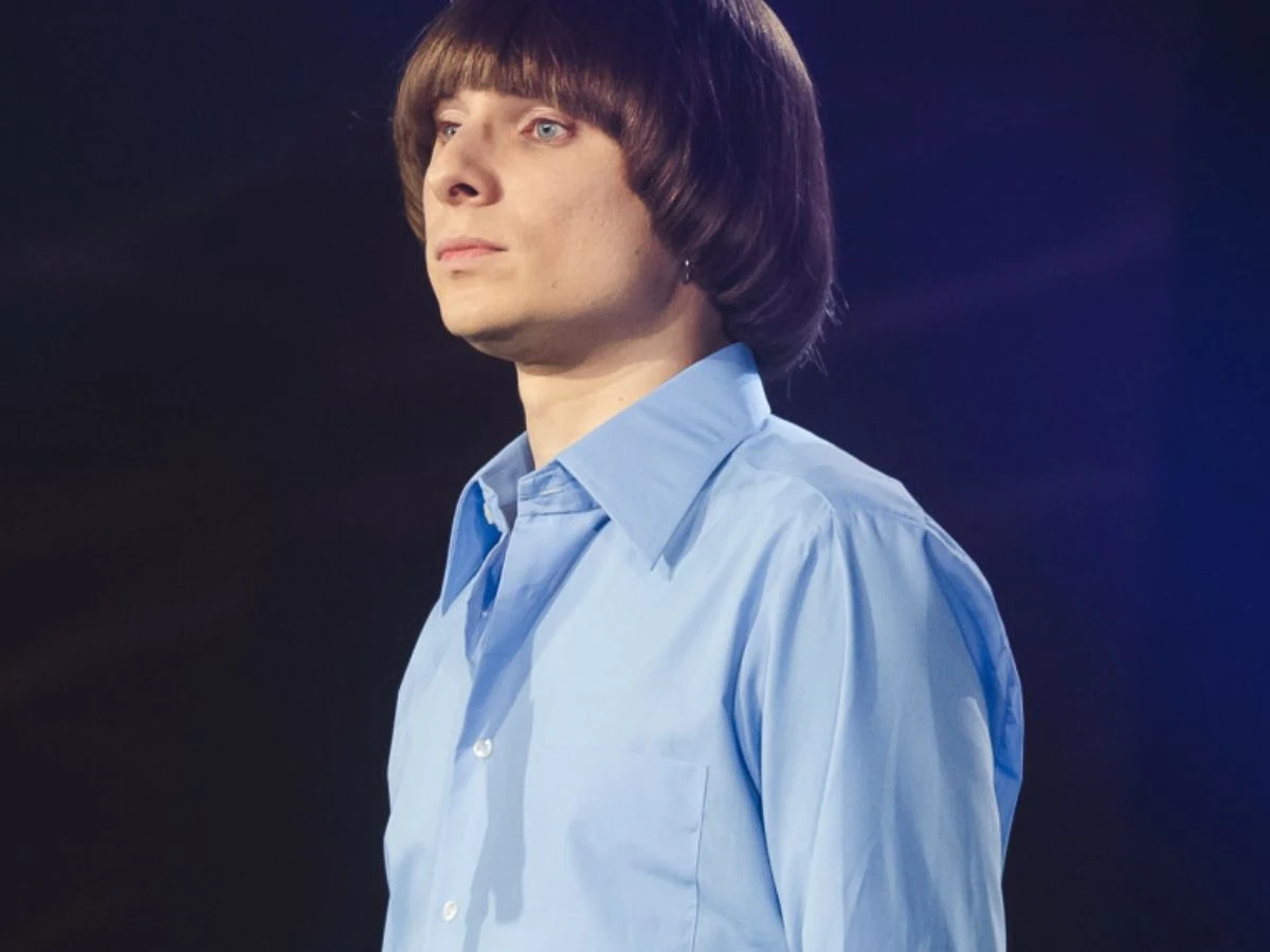 Ralph Kaminski Mężczyzna o jasnej cerze w niebieskiej koszuli z kołnierzykiem, z charakterystyczną fryzurą na tak zwanego „grzybka”, patrzy poważnie w bok na ciemnym tle.