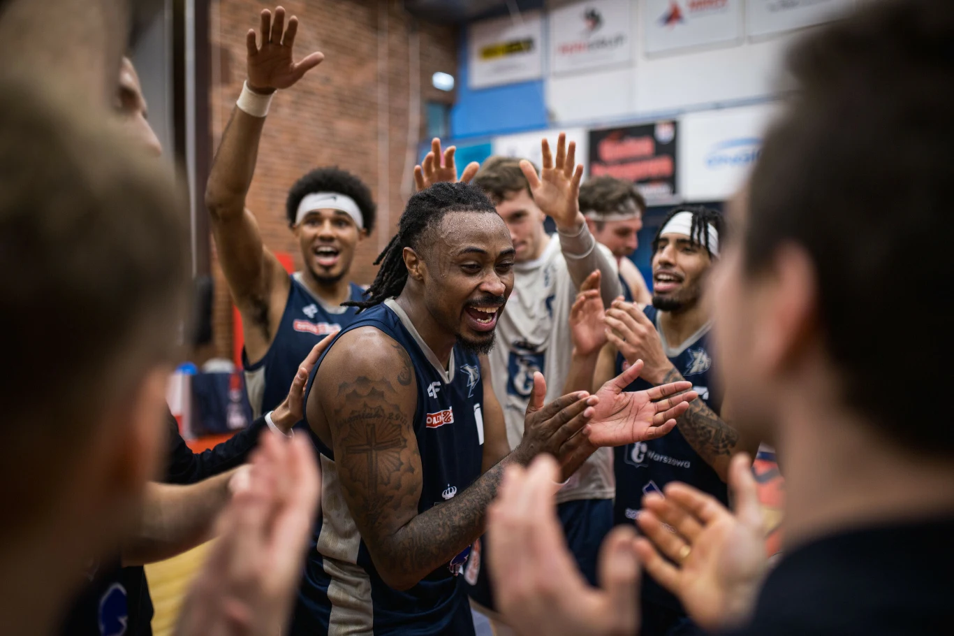 Koszykarze w sportowych strojach otoczeni przez kolegów z drużyny, unoszą ręce i klaszczą, uśmiechając się i okazując radość po zakończonym meczu. Na pierwszym planie centralna postać z dredami, otoczona przez pozostałych zawodników, w tle widoczne fra...