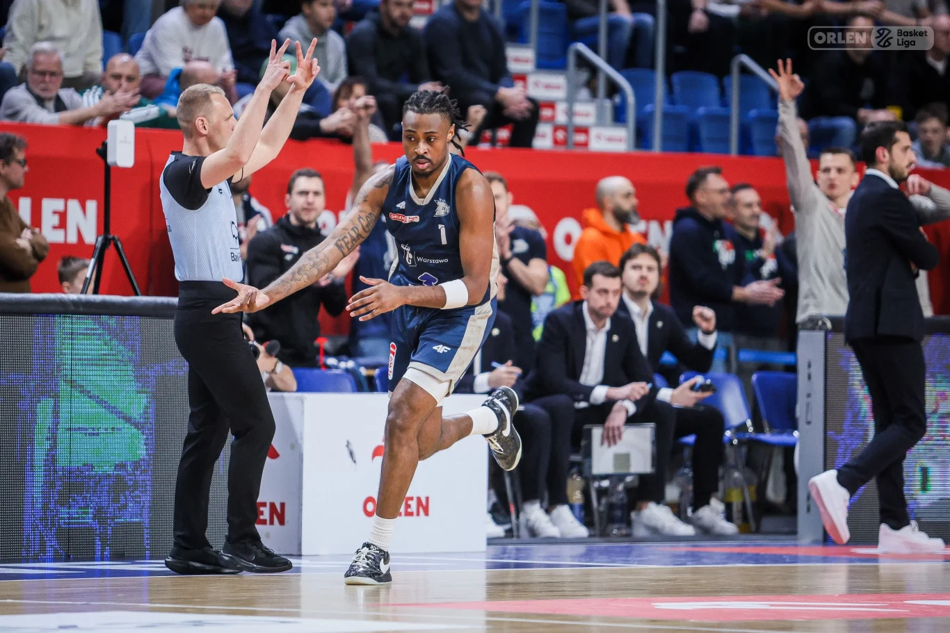 Zawodnik drużyny koszykarskiej biegnie po parkiecie, tuż obok stojącego sędziego sygnalizującego ręką decyzję. W tle widoczni kibice, ławka rezerwowych oraz trenerzy obserwujący przebieg meczu.