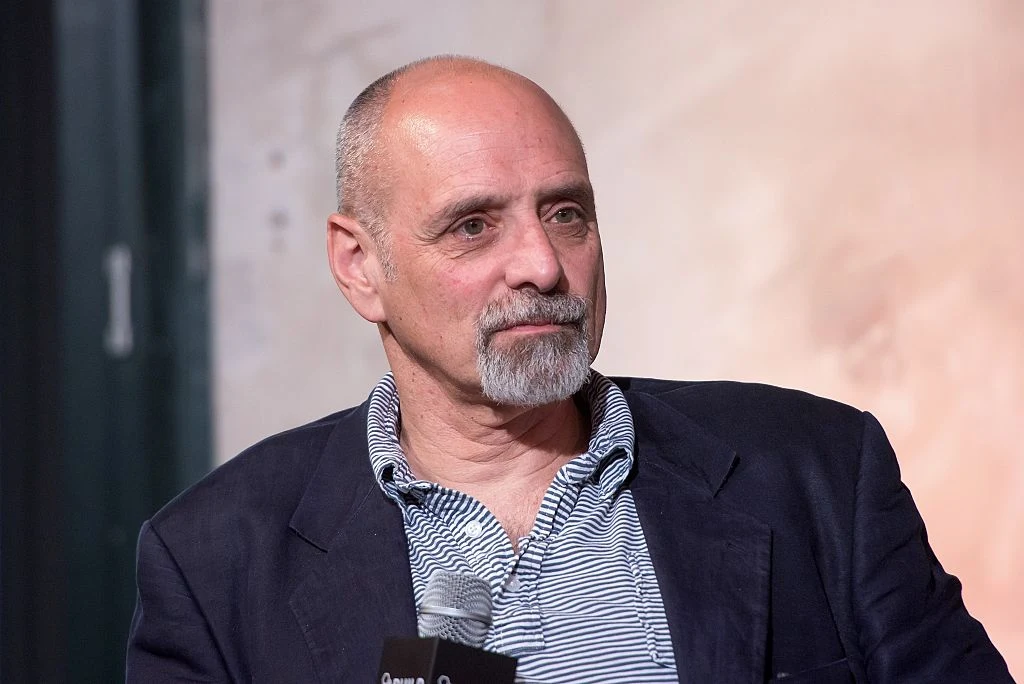 Eric Schlosser - autor tego artykułu i opisywanej książki Mężczyzna o siwych włosach i brodzie w marynarce oraz koszulce w paski, trzymający mikrofon i patrzący w bok, siedzi na tle rozmytego, jasnego tła.