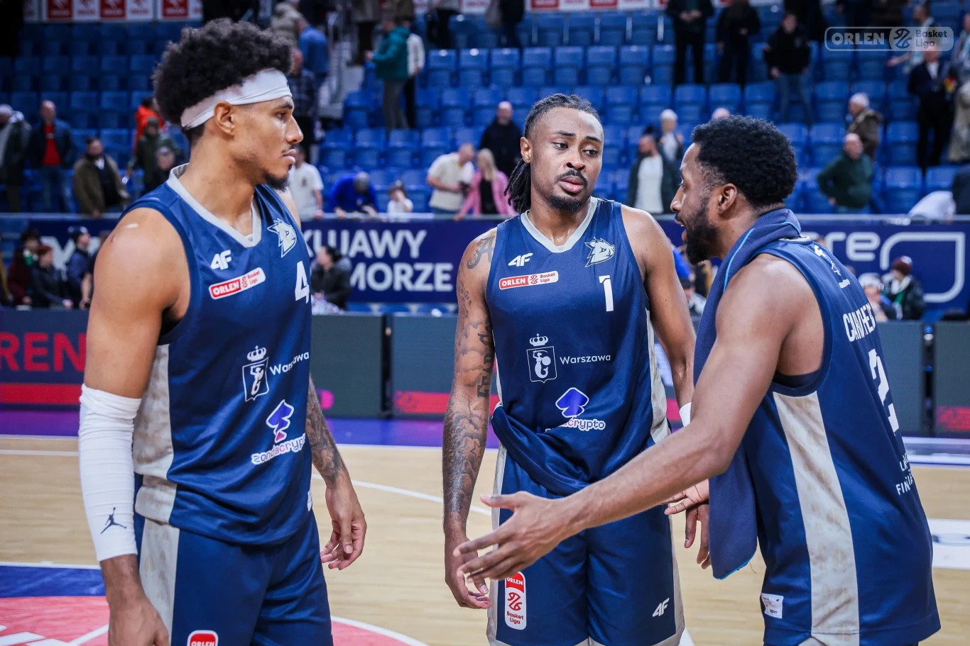 Trzech koszykarzy w niebiesko-białych strojach drużynowych prowadzi rozmowę na parkiecie hali sportowej, wokół widać trybuny z kibicami.