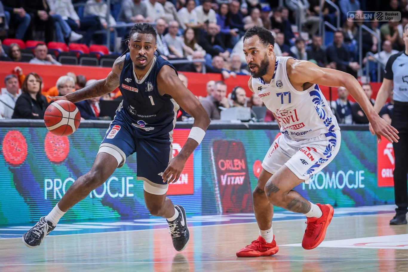 Dwóch koszykarzy w dynamicznej akcji podczas meczu, jeden zawodnik w granatowym stroju kozłuje piłkę, starając się minąć rywala w białym stroju, na trybunach widoczni kibice.