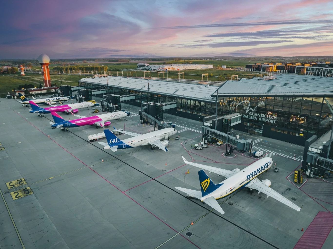 Samoloty pasażerskie różnych linii lotniczych ustawione przy terminalu lotniska podczas pięknego, kolorowego wschodu lub zachodu słońca. Nowoczesny terminal z dużymi przeszkleniami oraz pas startowy, w tle zieleń i dalsza zabudowa.