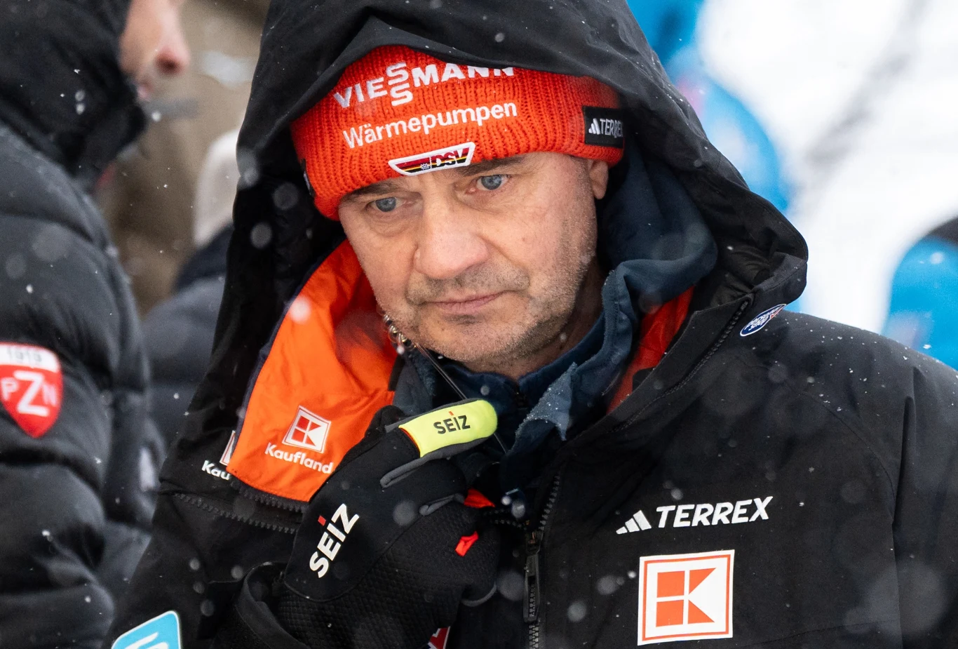 Stefan Horngacher Mężczyzna w zimowej kurtce i czerwonej czapce z logo Viessmann, trzymający mikrofon lub krótkofalówkę, otoczony przez śnieg i innych ludzi, najprawdopodobniej podczas wydarzenia sportowego na świeżym powietrzu.