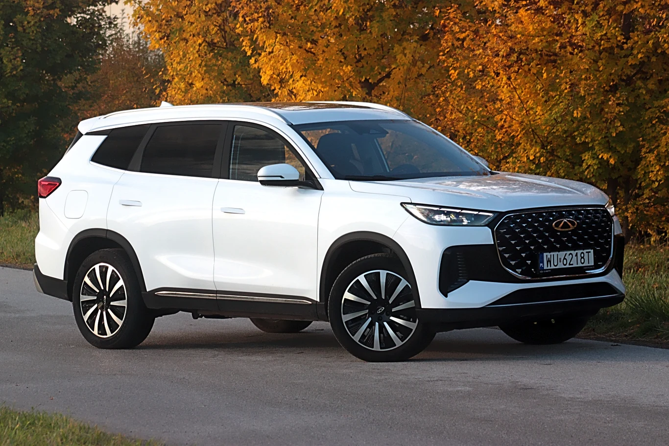 Biały nowoczesny SUV zaparkowany na niewielkiej uliczce na tle żółtych liści drzew, masywne felgi, elegancka linia nadwozia, ciemne szyby i charakterystyczny grill.