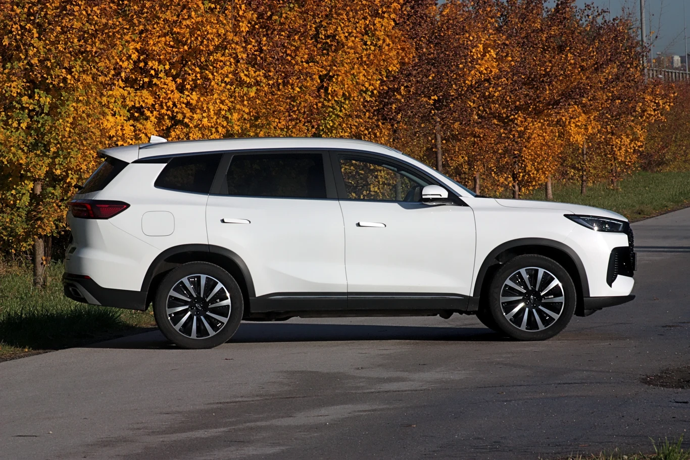 Biały SUV zaparkowany na bocznej, asfaltowej drodze, w tle kolorowe jesienne drzewa pokryte liśćmi w odcieniach żółci i pomarańczu.