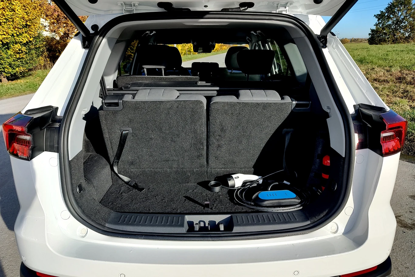 Otwarty bagażnik białego samochodu typu SUV zaparkowanego na drodze. W bagażniku widoczne przewody ładujące oraz czarne tapicerowane wnętrze, tylne siedzenia pozostają w pozycji pionowej.