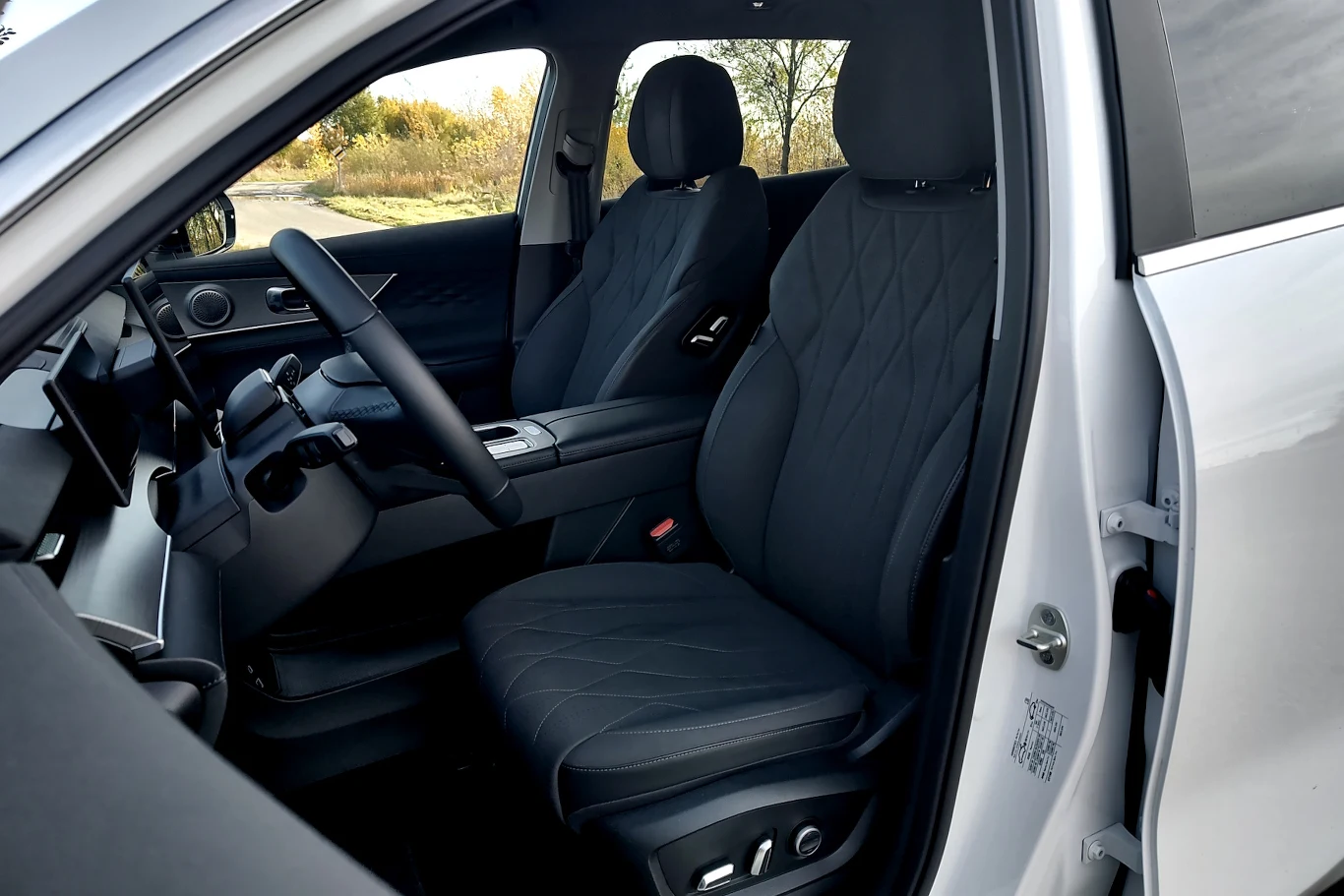 Nowoczesne wnętrze samochodu z ciemnymi tapicerowanymi fotelami, kierownicą po lewej stronie i konsolą centralną z widocznymi przyciskami. Na zewnątrz widoczna zieleń przez boczne okno.