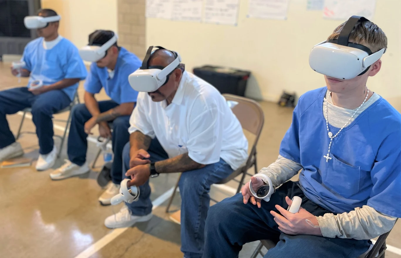 Wirtualna rzeczywistość pomaga więźniom przygotować się do powrotu do społeczeństwa Czterech mężczyzn siedzi na krzesłach, ubrani w niebieskie i białe ubrania, wszyscy korzystają z gogli wirtualnej rzeczywistości oraz kontrolerów trzymanych w rękach.