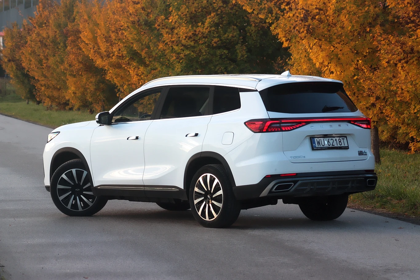 Biały SUV zaparkowany na poboczu asfaltowej drogi, w tle jesienne drzewa o żółto-pomarańczowych liściach, samochód ustawiony tyłem z widoczną tablicą rejestracyjną.