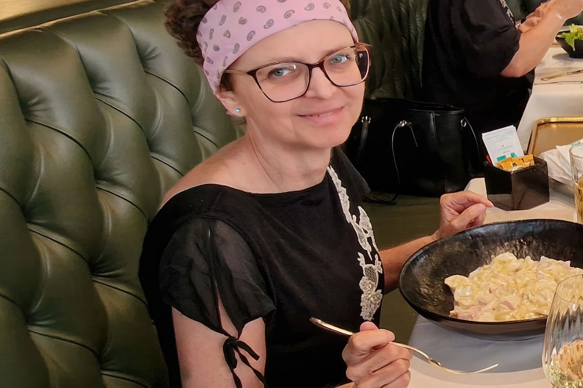 Kobieta w okularach i opasce na głowie siedzi przy stole w restauracji, spożywa makaron z czarnego talerza i uśmiecha się do obiektywu.