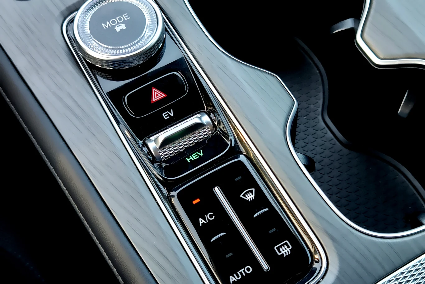 Panel centralny samochodu z widocznymi przyciskami do trybów jazdy EV i HEV, przyciskiem awaryjnym oraz sterowaniem klimatyzacją, wykonany z materiału imitującego drewno, z chromowanymi wykończeniami.