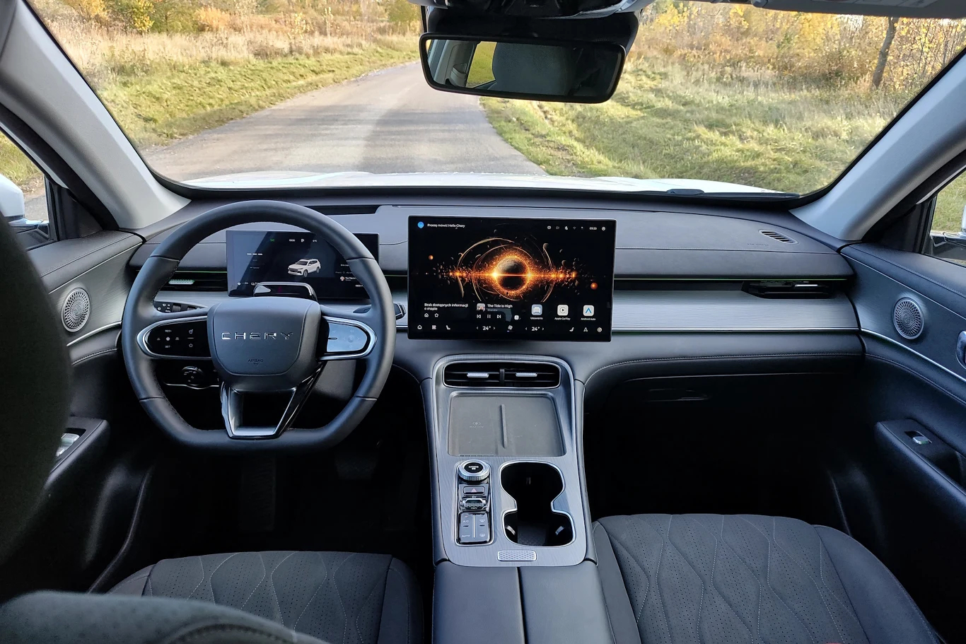 Nowoczesne wnętrze samochodu z dużym ekranem multimedialnym na konsoli środkowej, cyfrowymi zegarami i ergonomiczną kierownicą, widok przez przednią szybę na pustą, asfaltową drogę otoczoną zielenią.