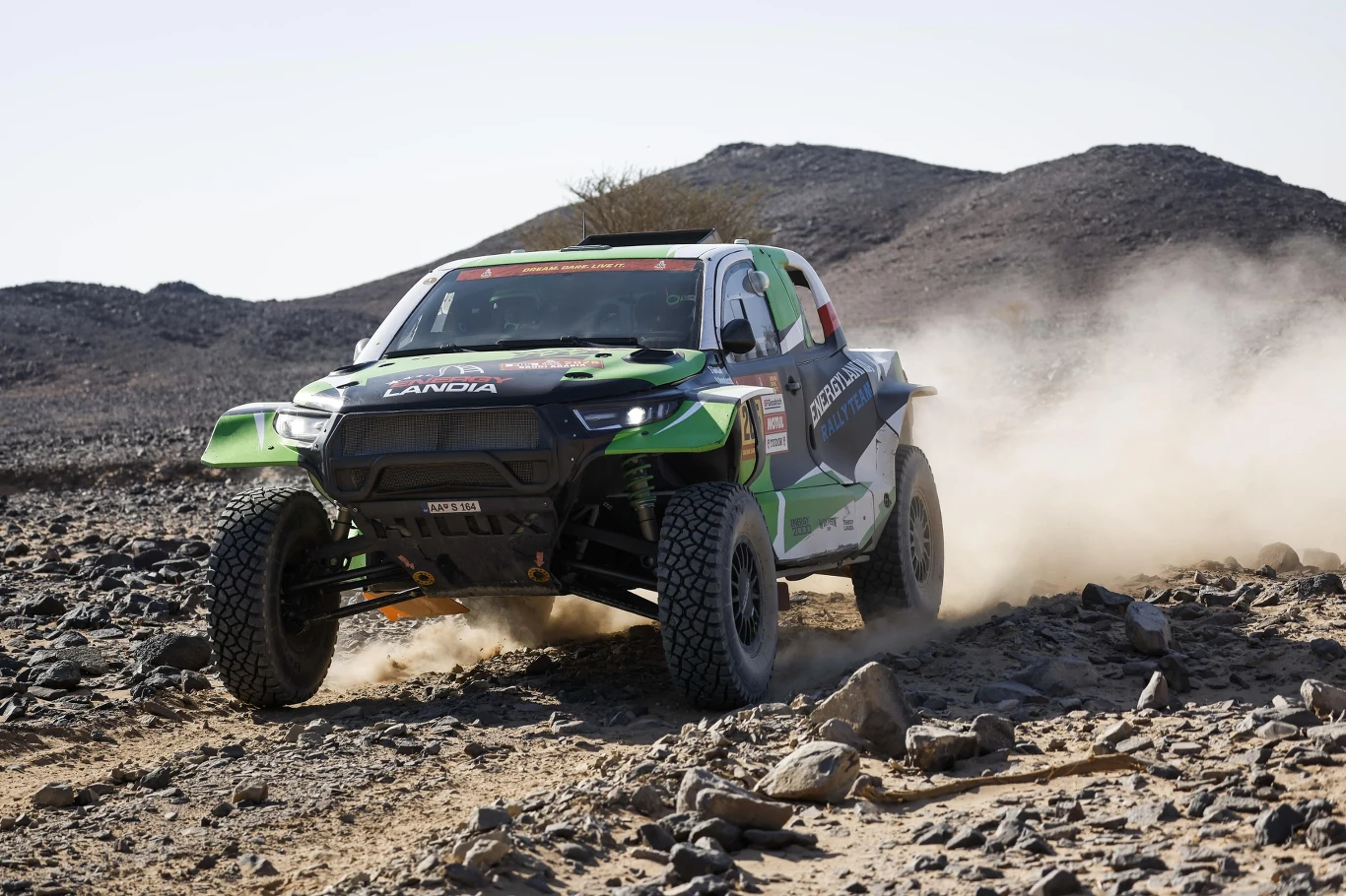 Samochód rajdowy pokonuje kamienistą, pustynną trasę, wzbijając za sobą kurz. W tle widoczne są niskie, skaliste wzgórza, a otoczenie jest surowe i suche.