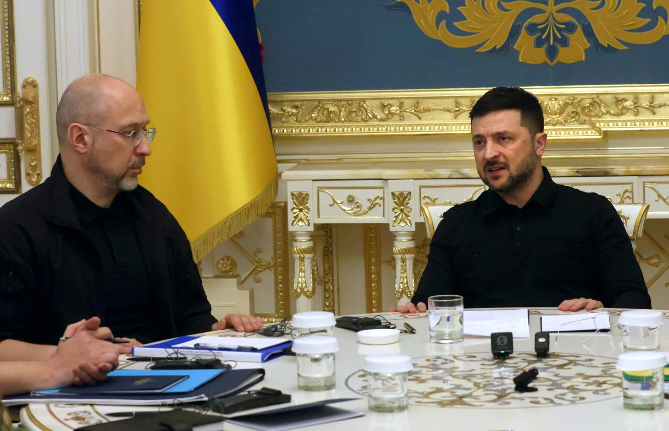 Zmiany w ukraińskim rządzie. Denys Szmyhal traci stanowisko ministra obrony Dwóch mężczyzn w ciemnych ubraniach siedzi przy okrągłym stole w eleganckim wnętrzu z bogato zdobionymi złotem ścianami. Przed nimi leżą dokumenty, notatniki i szklanki z wodą. Między nimi widać flagę narodową.