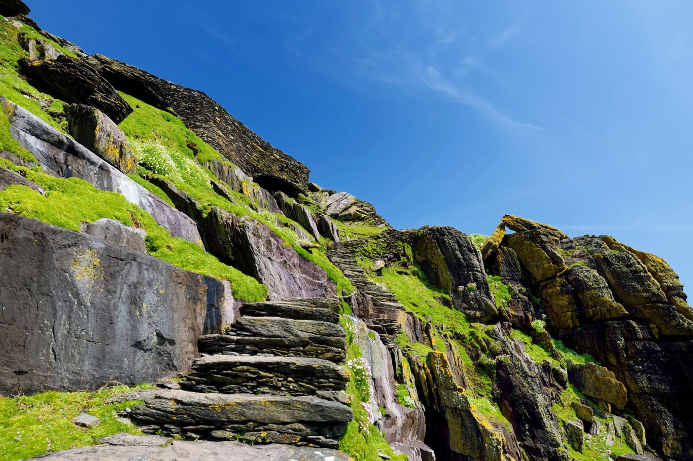 Kamienne schody wykute w skale prowadzą w górę pośród surowych, porośniętych zielonym mchem skał pod czystym, niebieskim niebem.