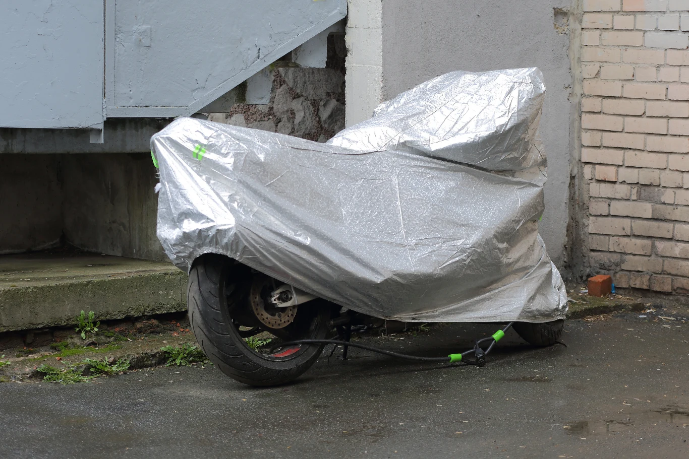 Jaki pokrowiec na motocykl wybrać? Motocykl przykryty srebrną plandeką, zabezpieczony łańcuchem na tylnej osi, stoi na mokrym asfalcie obok betonowego podestu przy ścianie z cegieł i tynku.