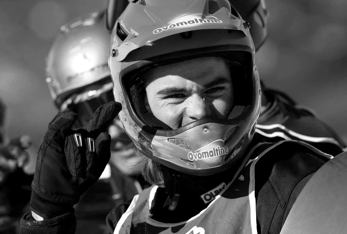 Młody zawodnik w kasku i stroju ochronnym uśmiecha się i wykonuje gest ręką, w tle widoczne są inne osoby również w kaskach.