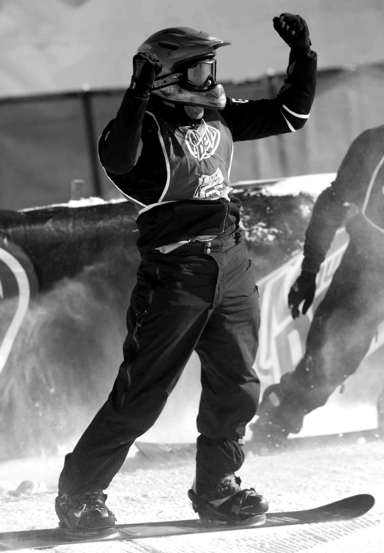 Osoba ubrana w sportową odzież i kask ochronny stoi triumfalnie na snowboardzie z uniesionymi rękami w geście zwycięstwa, w tle baner reklamowy oraz rozbryzgujący się śnieg.