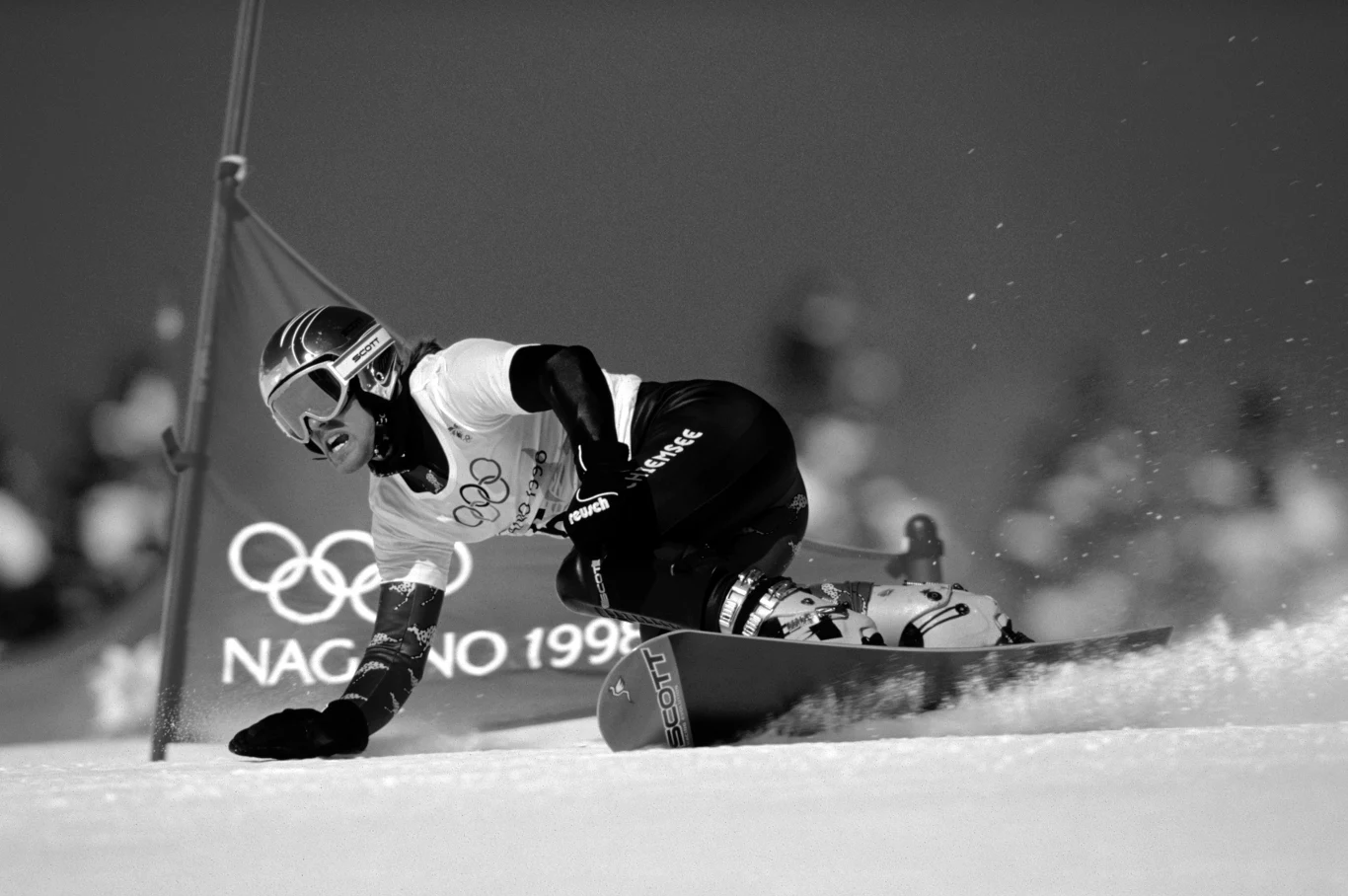 Zawodnik zjeżdżający na snowboardzie podczas zawodów olimpijskich w Nagano 1998, noszący kask oraz gogle ochronne, dynamiczny ruch przy pokonywaniu zakrętu na stoku, w tle widoczne rozmyte logo olimpijskie i napis Nagano 1998.