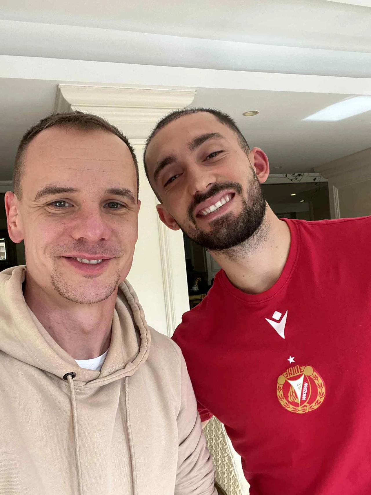Dwóch uśmiechniętych mężczyzn pozuje razem do selfie w jasnym wnętrzu. Jeden z nich ma na sobie jasną bluzę z kapturem, a drugi czerwony sportowy t-shirt z herbem klubu piłkarskiego.