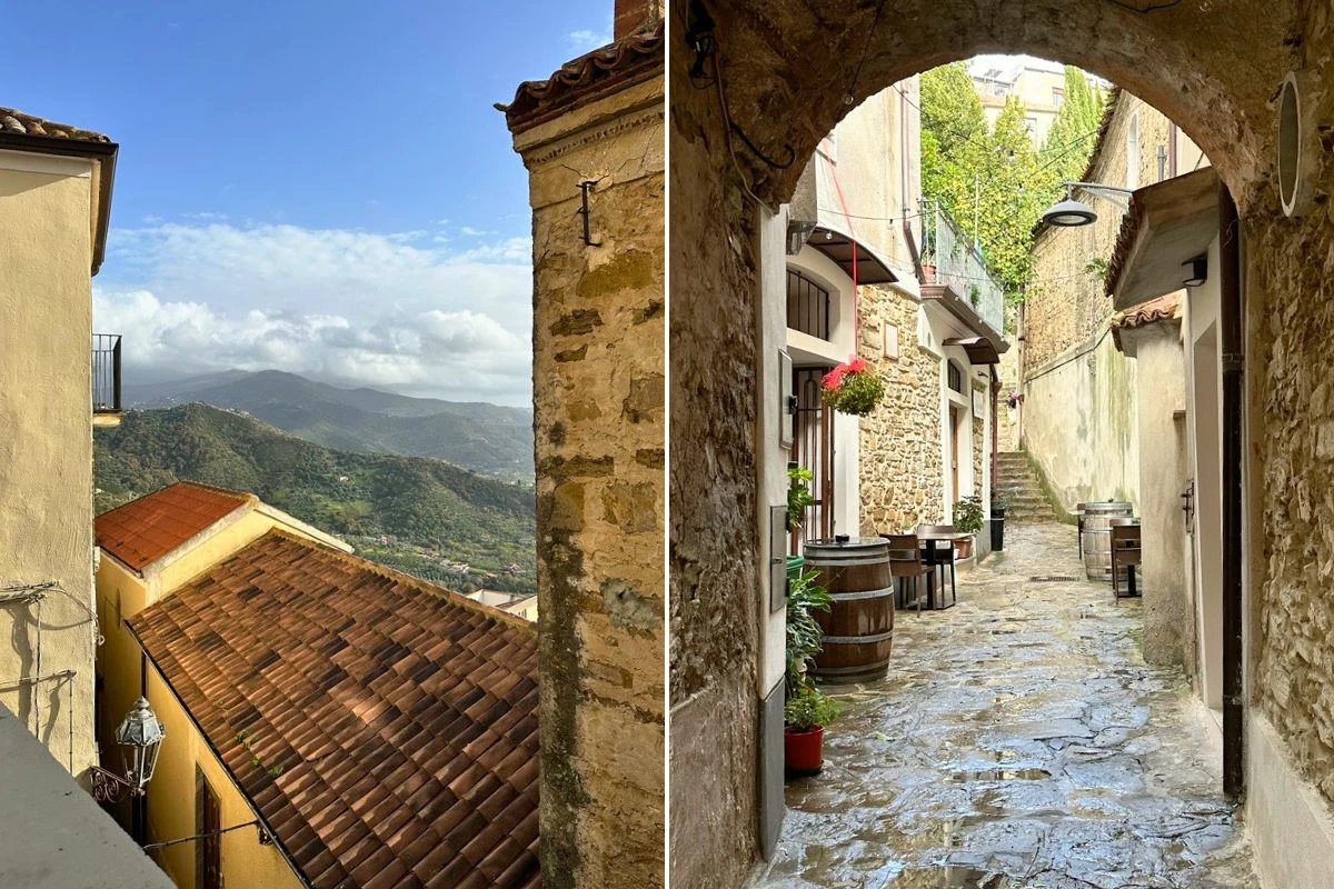 Widok na stare budynki o ceglastych dachach, góry w oddali pod błękitnym niebem oraz wąska, brukowana uliczka z kwiatami, beczkami i detalami architektonicznymi, otoczona ścianami kamienic.