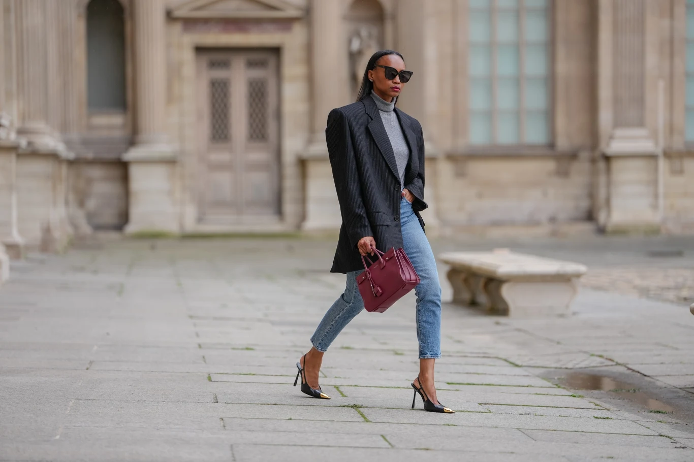 Elegancka kobieta w okularach przeciwsłonecznych ubrana w czarną marynarkę, szary golf, niebieskie jeansy i czarne szpilki idzie po kamiennym dziedzińcu trzymając bordową torebkę, w tle zabytkowy budynek.