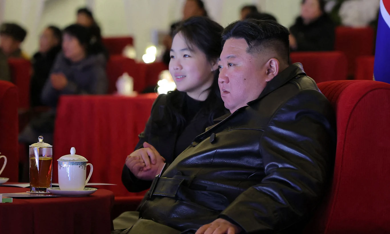 Mężczyzna w czarnym skórzanym płaszczu siedzi obok kobiety na czerwonym fotelu w eleganckim otoczeniu, na stoliku obok stoi filiżanka z herbatą pod przykryciem i szklanka z napojem. W tle widoczni są inni uczestnicy wydarzenia.