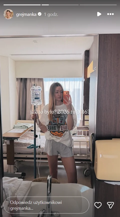 Młoda kobieta robi sobie zdjęcie w lustrze stojąc w szpitalnym pokoju obok łóżka szpitalnego i stojaka do kroplówki, ubrana w luźny t-shirt oraz krótkie spodenki, w tle widać jasne zasłony i część sprzętu medycznego.