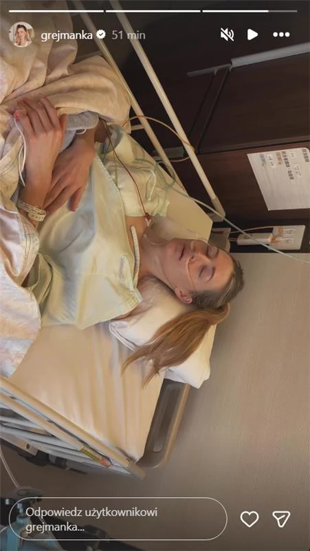 Kobieta leży w szpitalnym łóżku, ubrana w szpitalną koszulę, z założonym wenflonem na ręce, z zamkniętymi oczami. W tle widoczny jest fragment szpitalnego sprzętu i szafka na dokumenty.