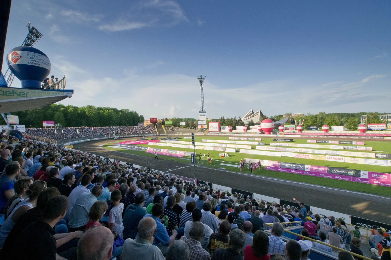Tłumy kibiców zgromadzone na trybunach stadionu podczas dużego wydarzenia sportowego, kolorowe banery reklamowe wokół toru żużlowego, jasno oświetlone niebo oraz infrastruktura stadionowa.