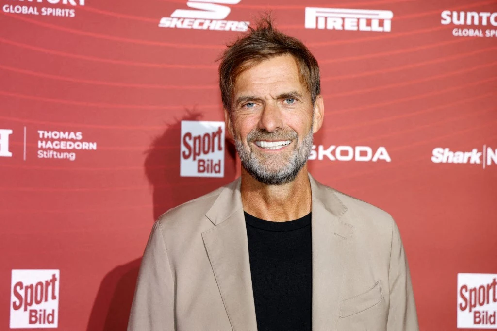 Uśmiechnięty mężczyzna z zarostem w jasnej marynarce i czarnej koszulce stoi na tle ścianki promocyjnej z logotypami różnych marek.