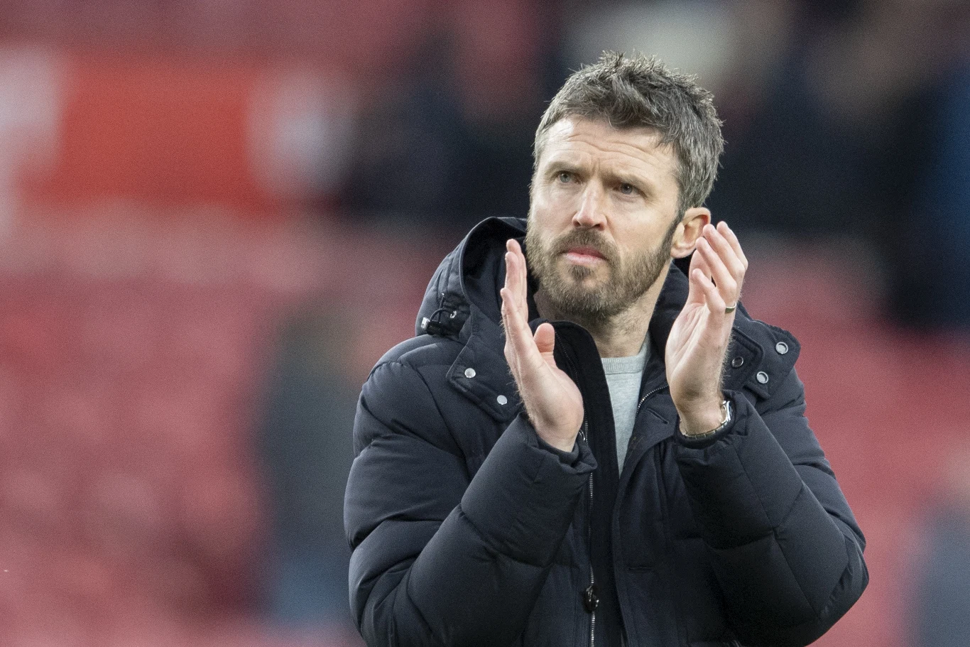 Mężczyzna z zarostem ubrany w ciemną kurtkę stoi na tle rozmytego stadionu i klaszcze dłońmi, sprawiając wrażenie skoncentrowanego.