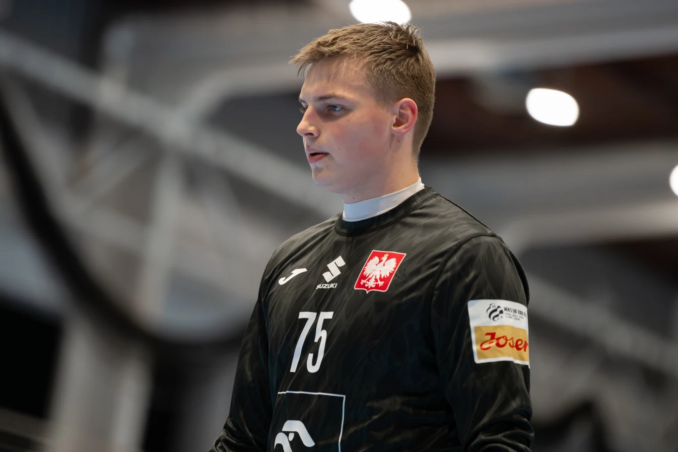 Młody mężczyzna w czarnym stroju sportowym z numerem 75 i godłem Polski, skoncentrowany, w hali sportowej podczas wydarzenia.