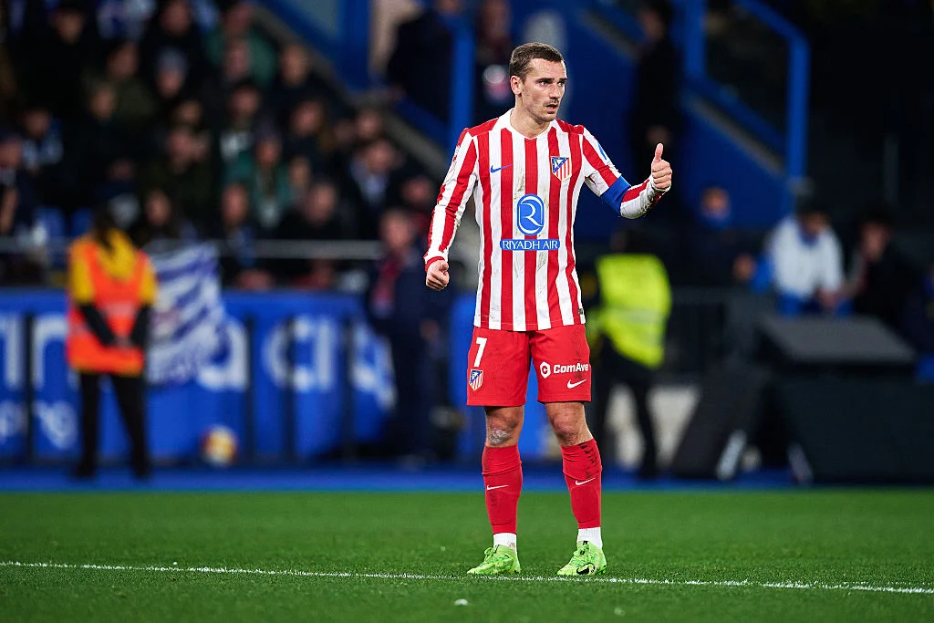 Piłkarz w stroju Atlético Madryt stoi na murawie stadionu, unosi kciuk w geście aprobaty, w tle widać nieostre sylwetki kibiców i obsługi technicznej.