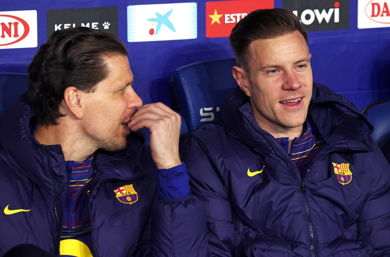 Dwóch mężczyzn w kurtkach z herbem FC Barcelona siedzi obok siebie na ławce rezerwowych. Jeden z nich rozmawia z drugim, obaj wydają się zrelaksowani.