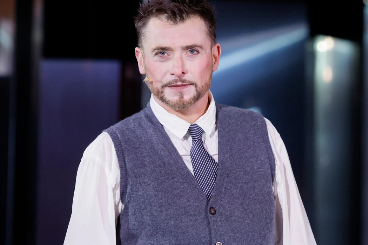 Kacper Kuszewski Mężczyzna z zarostem ubrany w elegancką białą koszulę, szary kamizelkę oraz krawat, stoi na tle rozmytego wnętrza z jasnym światłem w tle.