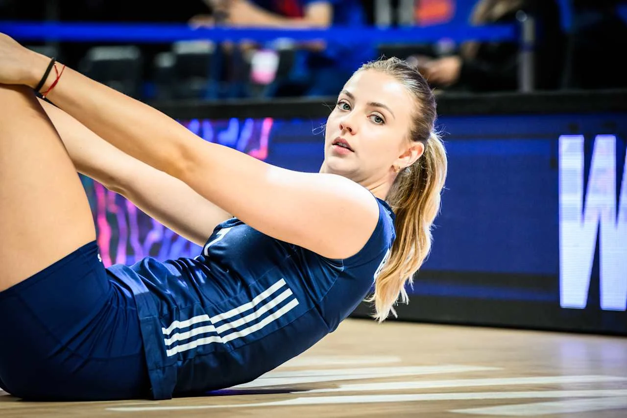 Kobieta w sportowym stroju wykonuje ćwiczenie na podłodze hali sportowej, jej dłonie wsparte są na nogach, na twarzy wyraz koncentracji, w tle widoczne elementy wyposażenia hali i bariera z niebieską taśmą.