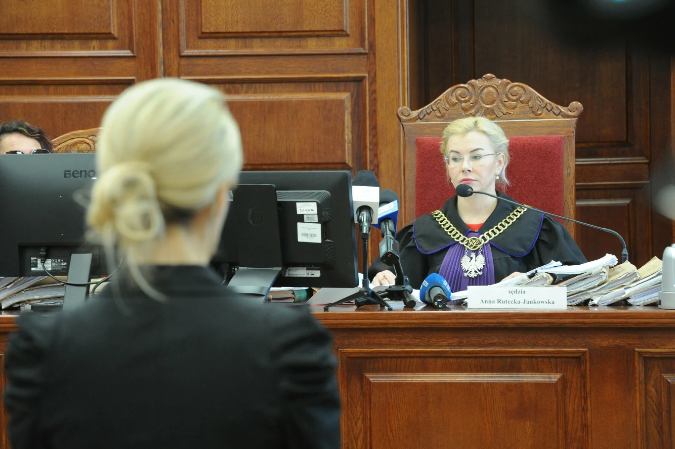 Koszalin. Ponownie rusza proces "Krwawego Tulipana". Na zdjęciu sędzia Anna Rutecka-Jankowska Sędzia w todze siedząca za dużym, drewnianym biurkiem w sali sądowej, przed nią stos dokumentów i mikrofony. Na pierwszym planie kobieta z blond włosami zwrócona plecami do aparatu.