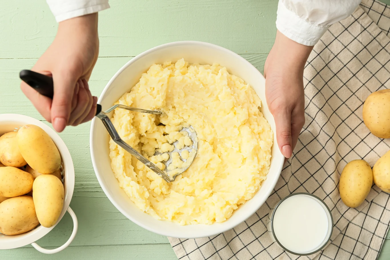 Ręce trzymające tłuczek do ziemniaków w trakcie robienia puree, miska z ugotowanymi i ugniecionymi ziemniakami, świeże ziemniaki po lewej stronie oraz szklanka mleka na kuchennej ściereczce.
