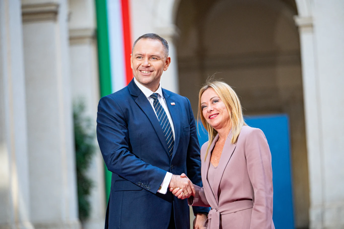 Dwójka elegancko ubranych osób podaje sobie ręce, uśmiechając się do aparatu. W tle widoczna kolumna budynku i pionowy pas flagi w barwach zieleni, bieli i czerwieni.