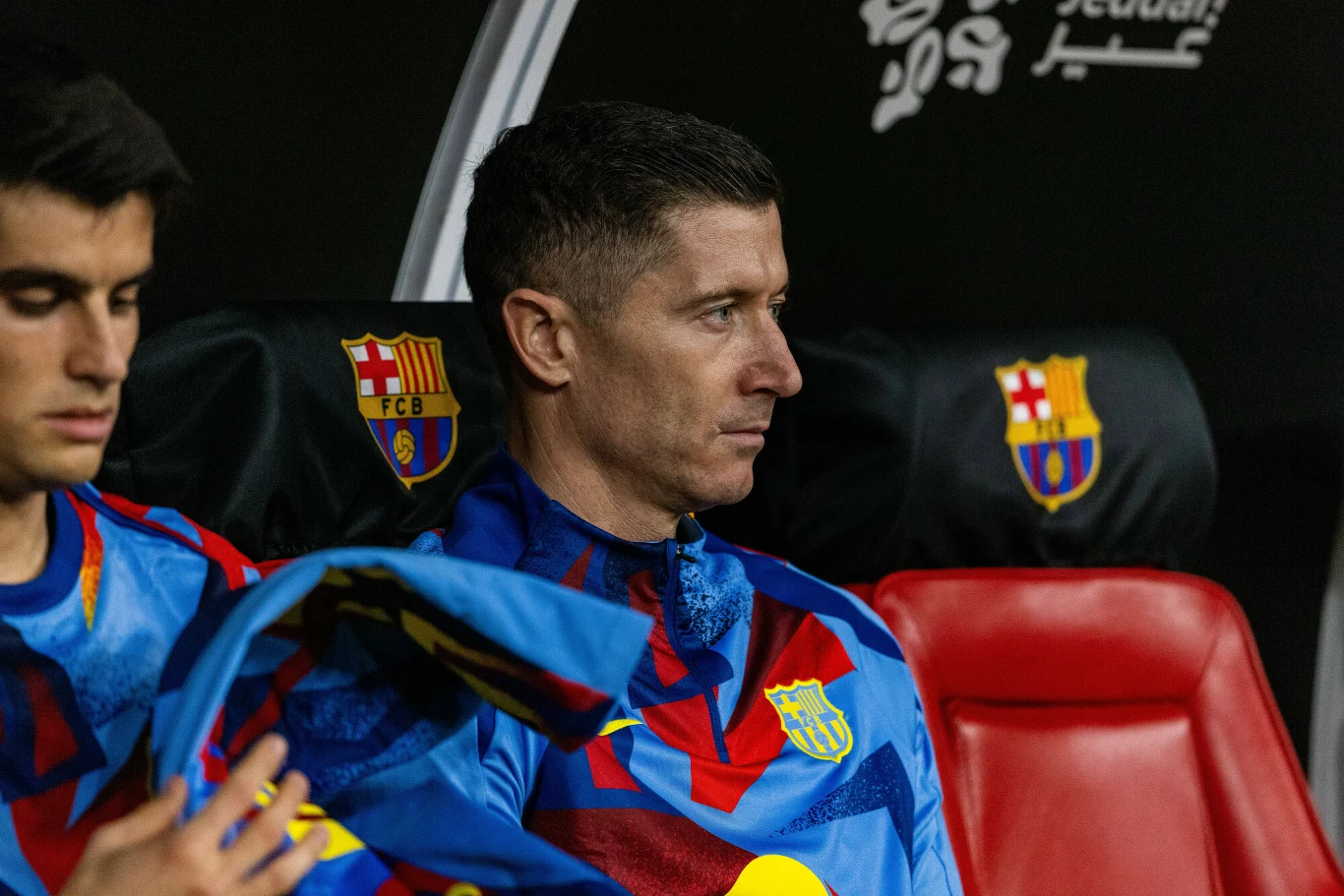Robert Lewandowski w FC Barcelona Piłkarz FC Barcelony w stroju klubowym siedzi na ławce rezerwowych, z profilu, obok inny zawodnik trzymający koszulkę.